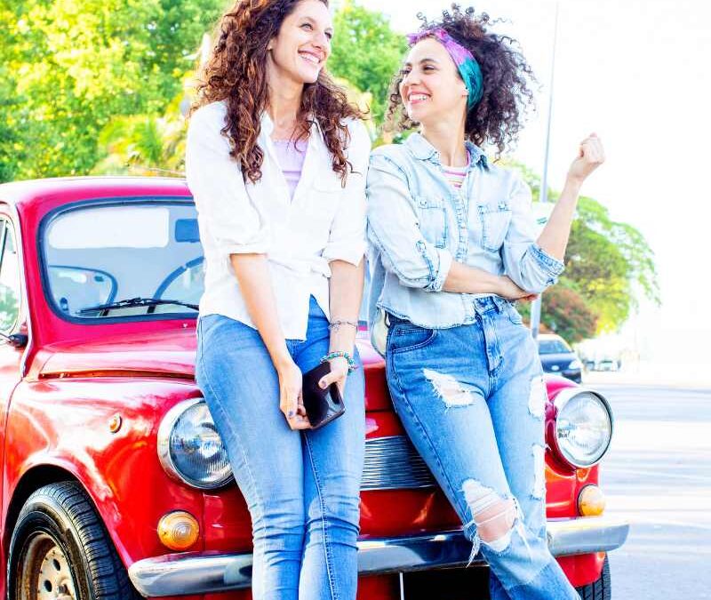 Two women in casual outfits smile and lean against the front of a vintage red car parked outdoors on a sunny day - thebestalbioninsuranceagent.com