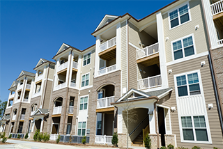 A modern, multi-story apartment building with tan siding, brick accents, and balconies with white railings under a clear blue sky - www.thebestalbioninsuranceagent.com